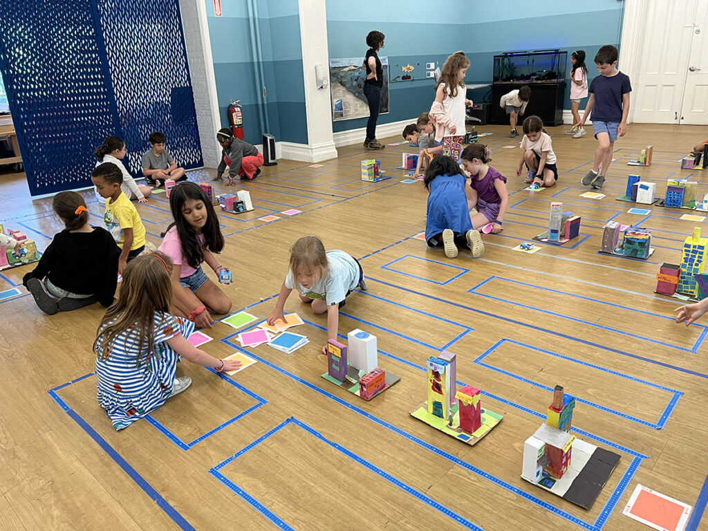 Students sit on the floor among paper versions of buildings. 