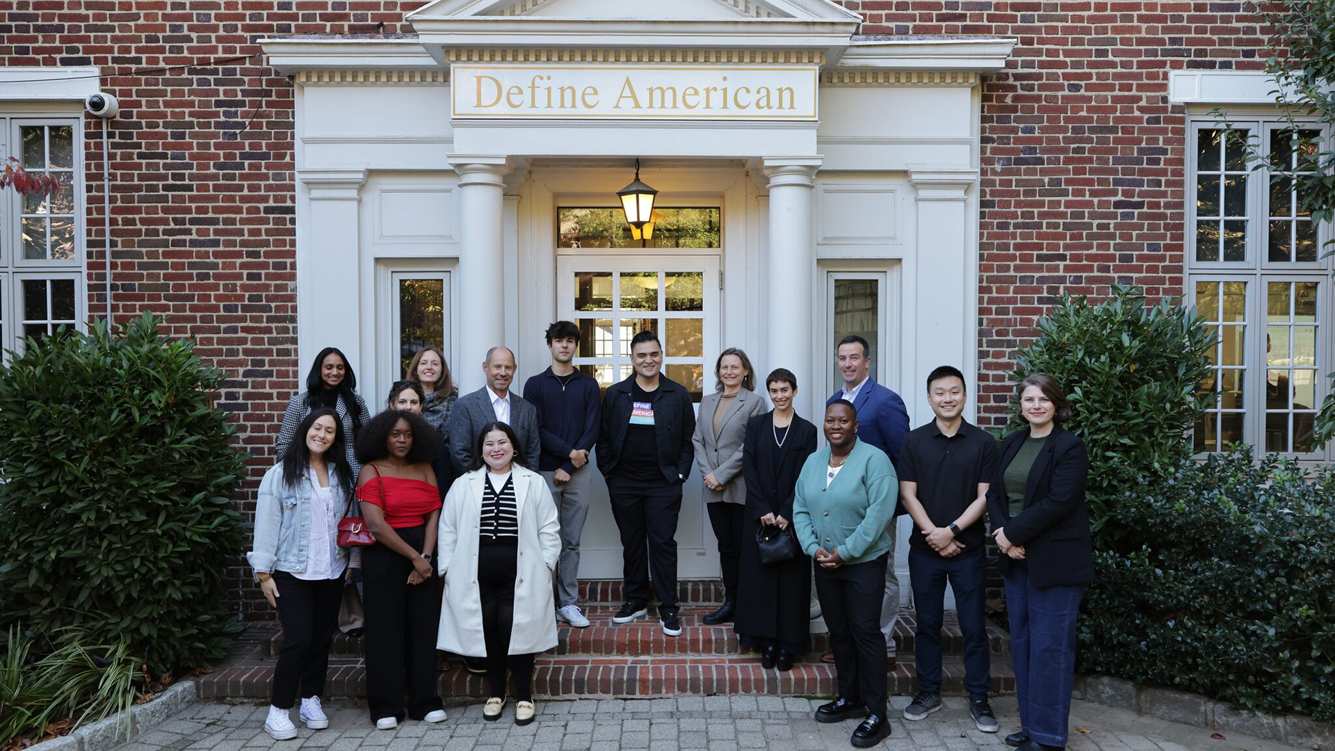 A group of people stand in front of a building with a sign that reads "Define American." 